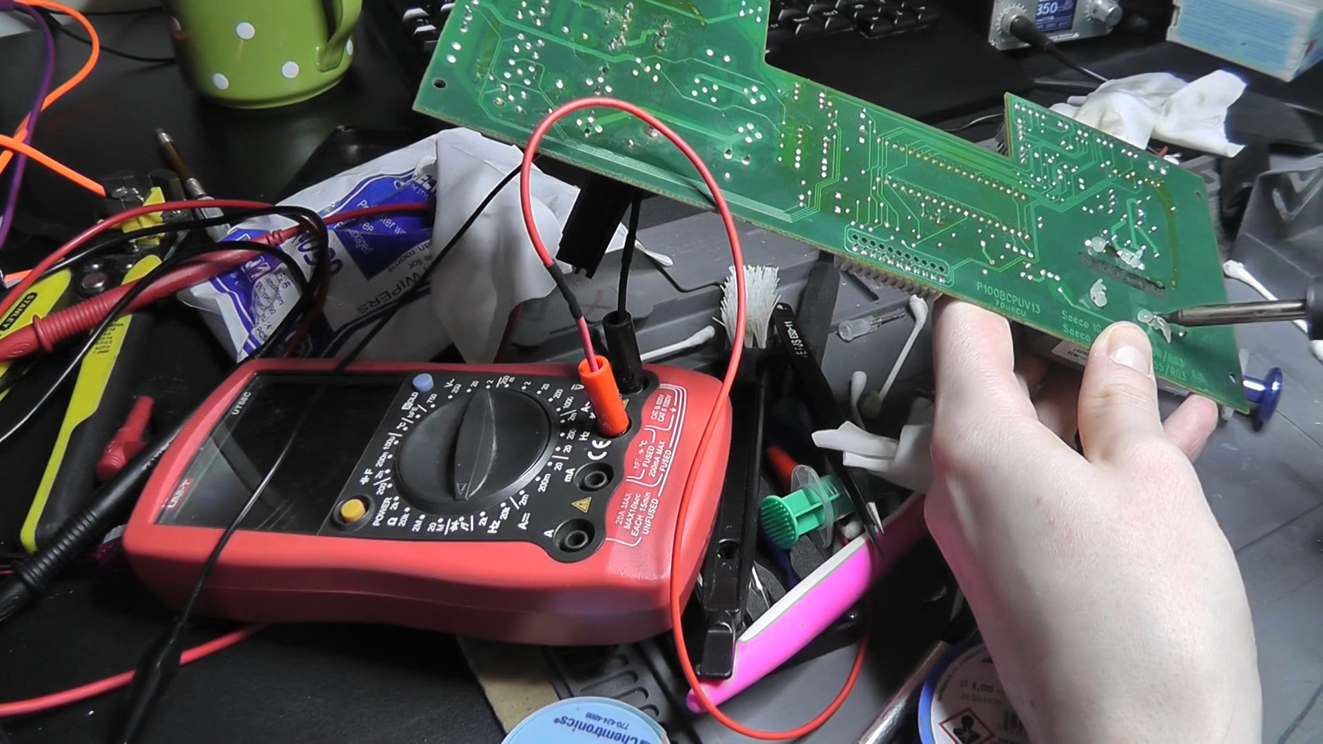 Lutowanie elementu na zielonej płycie drukowanej urządzenia elektronicznego, z użyciem czerwonego multimetru Unit-T, na zagraconym stole warsztatowym z widocznymi narzędziami i akcesoriami.