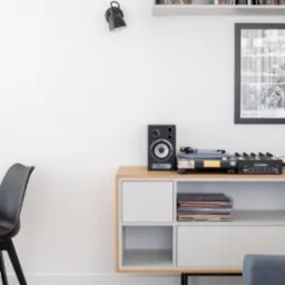 Minimalistyczny salon z gramofonem, głośnikiem, mikserem, płytami winylowymi na komodzie i półkach, czarną lampą na ścianie, plakatem w ramie, słuchawkami i książką na białym stoliku kawowym...