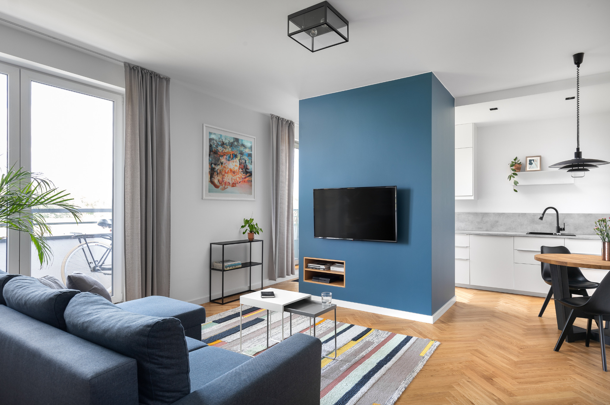 Nowoczesny salon z aneksem kuchennym w Warszawie. Niebieska ściana z telewizorem, szara sofa, stoliki kawowe, dywan w pasy, podłoga w jodełkę, biała kuchnia z czarną armaturą.
