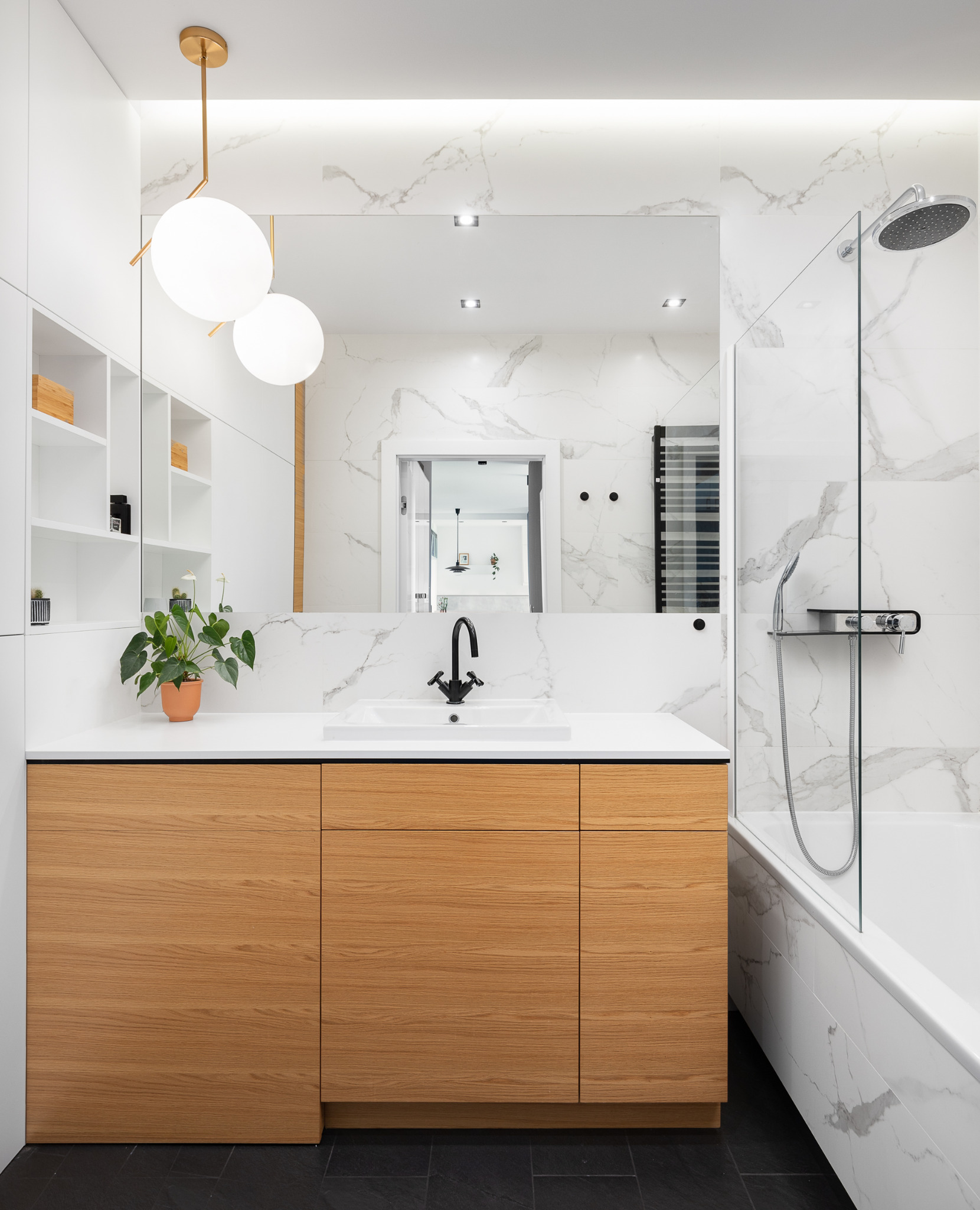 Nowoczesna łazienka w bieli i drewnie, z marmurowymi płytkami, czarną armaturą i okrągłymi lampami wiszącymi, widok na lustro odbijające dalszą część domu.