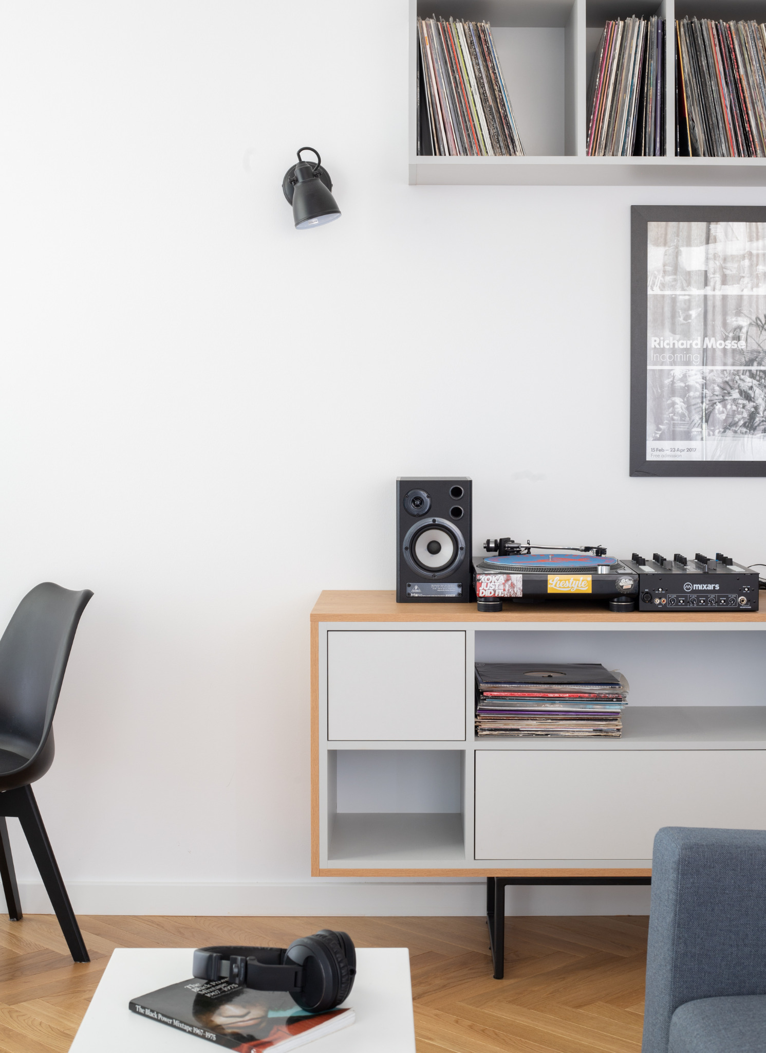 Minimalistyczny salon z gramofonem, głośnikiem, mikserem, płytami winylowymi na komodzie i półkach, czarną lampą na ścianie, plakatem w ramie, słuchawkami i książką na białym stoliku kawowym...