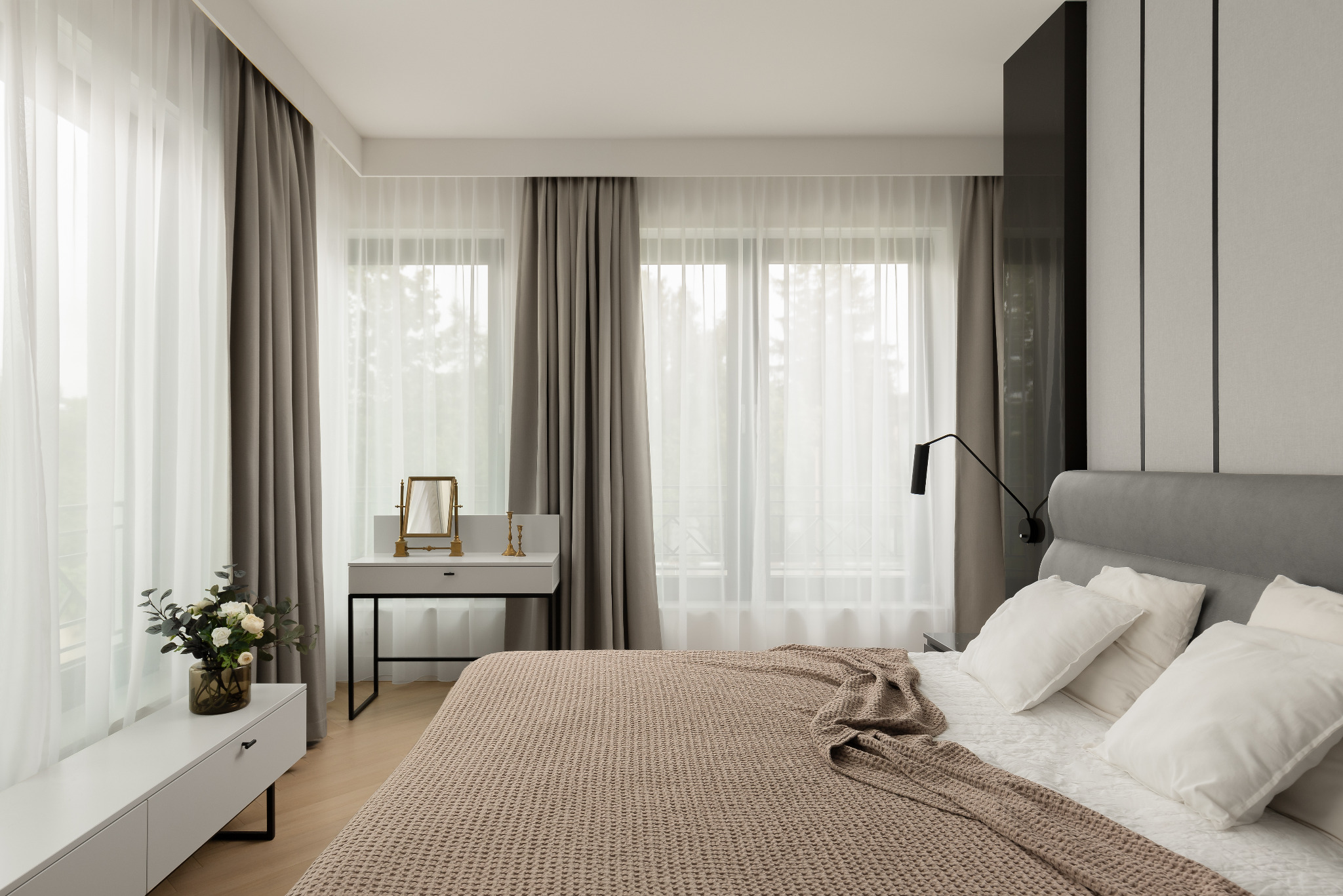 Jasna sypialnia z dużym oknem, zasłoniętym przezroczystymi i szarymi zasłonami, łóżko z beżową narzutą w wafelkowym wzorze, białe poduszki, toaletka z lustrem i bukiet kwiatów na szafce nocnej.