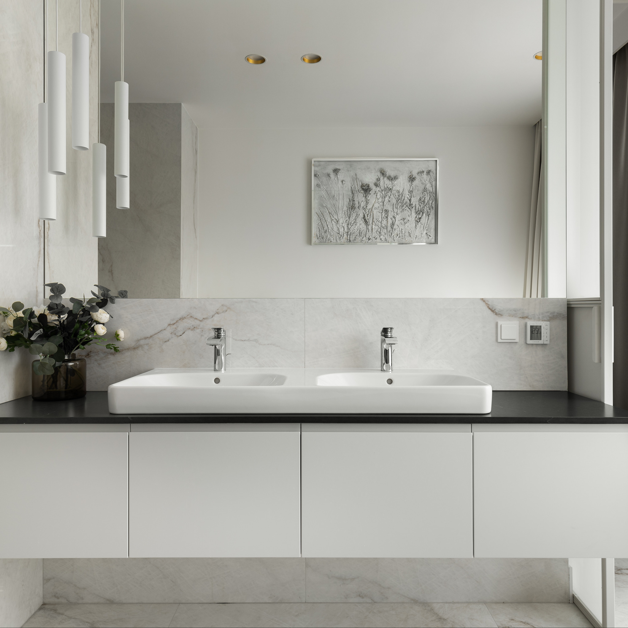 Nowoczesna łazienka z podwójną umywalką nablatową, czarnym blatem, białymi szafkami i marmurowym wykończeniem ściany, oświetlona wiszącymi lampami cylindrycznymi.