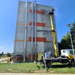 ODA SPÓŁKA Z OGRANICZONĄ ODPOWIEDZIALNOŚCIĄ - Żółty podnośnik koszowy na tle wysokiego budynku szpitalnego z rzędami kominów i częściowo odnowioną elewacją, widok z dołu na błękitne niebo.