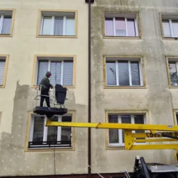 Pracownik w koszu podnośnika teleskopowego, na tle elewacji budynku w trakcie czyszczenia; widoczna różnica między umytą, jasną powierzchnią a brudną, ciemniejszą częścią ściany.