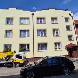 ODA SPÓŁKA Z OGRANICZONĄ ODPOWIEDZIALNOŚCIĄ - Żółta elewacja trzypiętrowego budynku z białymi oknami, widoczny żółty podnośnik na samochodzie dostawczym i czarny samochód osobowy na pierwszym planie.