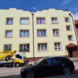 Żółta elewacja trzypiętrowego budynku z białymi oknami, widoczny żółty podnośnik na samochodzie dostawczym i czarny samochód osobowy na pierwszym planie.