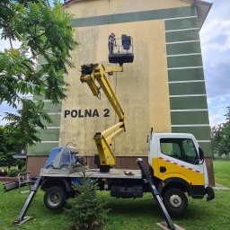 ODA SPÓŁKA Z OGRANICZONĄ ODPOWIEDZIALNOŚCIĄ - Pracownik w czerwonym kasku w koszu ruchomego podnośnika na tle budynku z napisem POLNA 2; żółty wysięgnik, biało-żółta ciężarówka na trawniku.