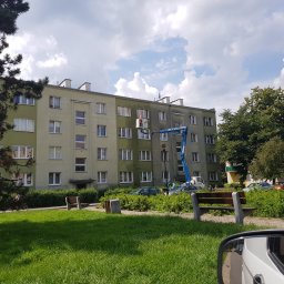 ODA SPÓŁKA Z OGRANICZONĄ ODPOWIEDZIALNOŚCIĄ - Pracownik w pomarańczowym kasku na podnośniku koszowym podczas prac na elewacji budynku wielorodzinnego, widok zza drzewa i fragmentu lusterka samochodu.