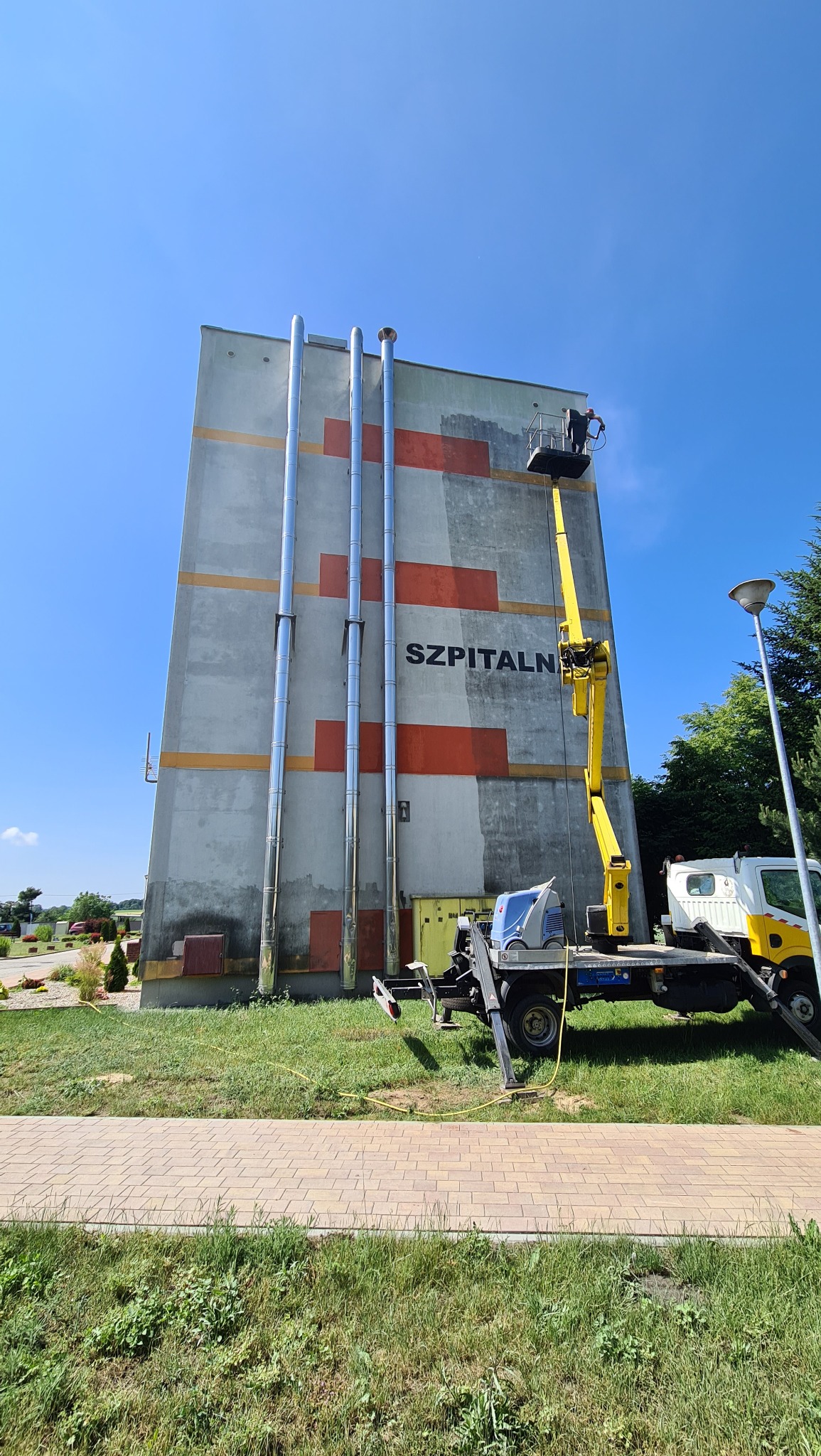 Żółty podnośnik koszowy na tle wysokiego budynku szpitalnego z rzędami kominów i częściowo odnowioną elewacją, widok z dołu na błękitne niebo.