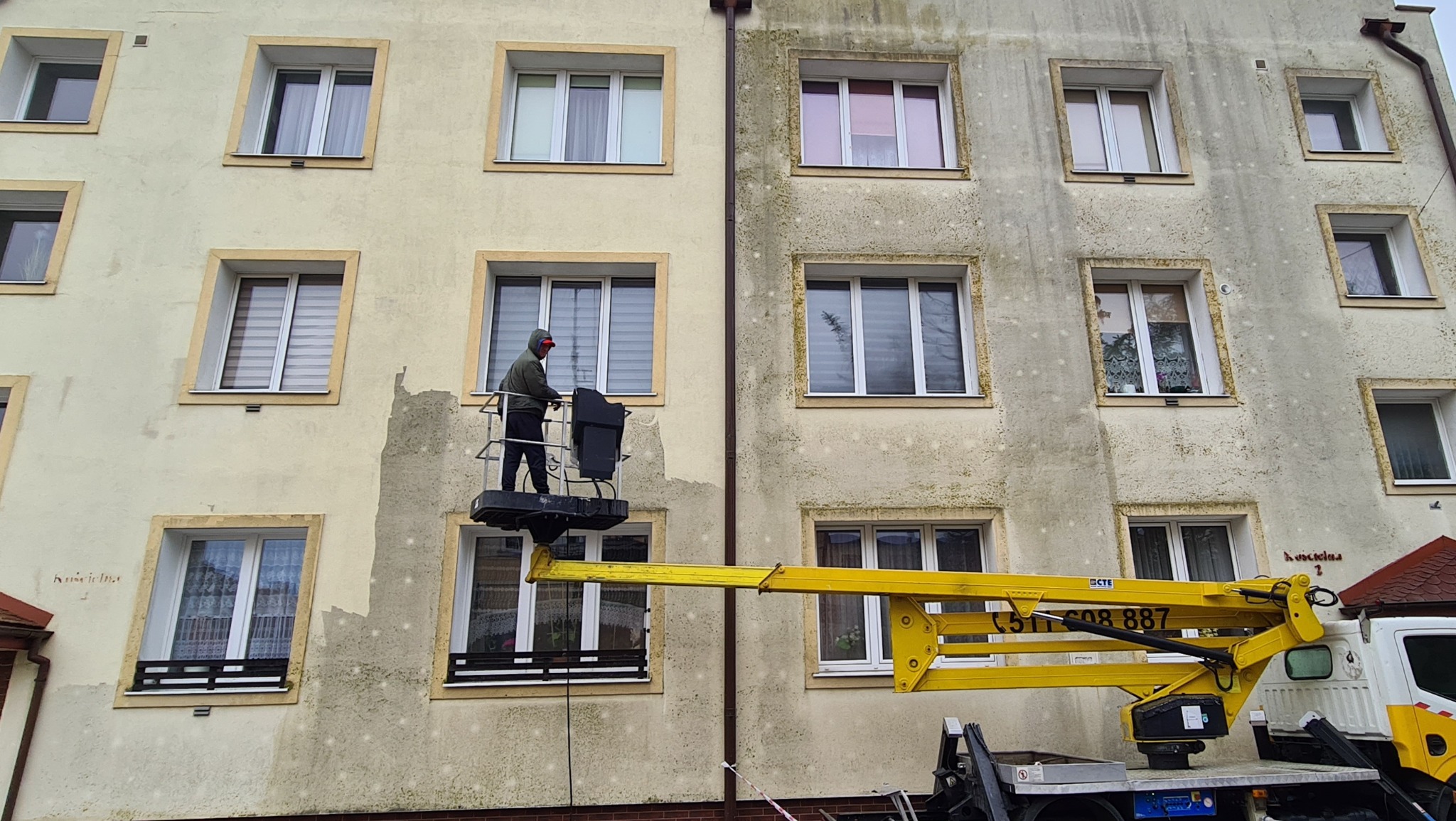 Pracownik w koszu podnośnika teleskopowego, na tle elewacji budynku w trakcie czyszczenia; widoczna różnica między umytą, jasną powierzchnią a brudną, ciemniejszą częścią ściany.