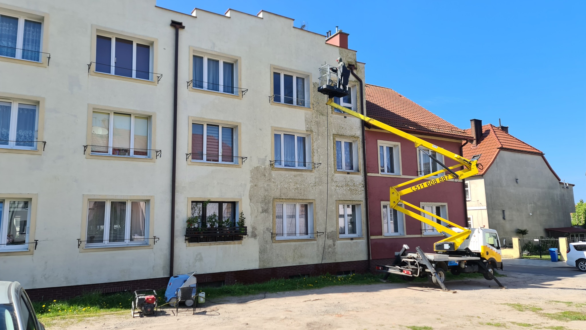Żółty podnośnik koszowy ustawiony przy bloku mieszkalnym, robotnik w koszu czyści elewację, widoczna różnica w kolorze między umytą a brudną częścią ściany.