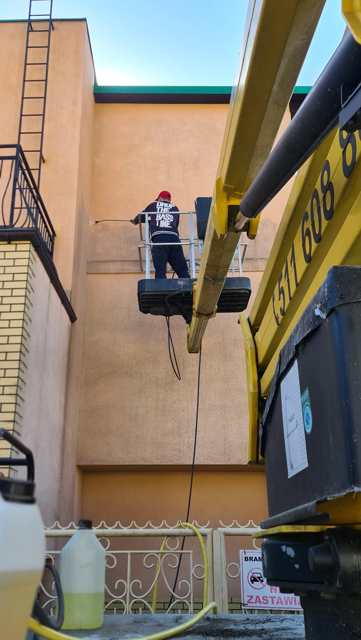 Pracownik w czerwonej czapce, stojący w koszu podnośnika, spryskuje elewację budynku pomarańczową cieczą z węża pod ciśnieniem, widok z dołu na tle błękitnego nieba.