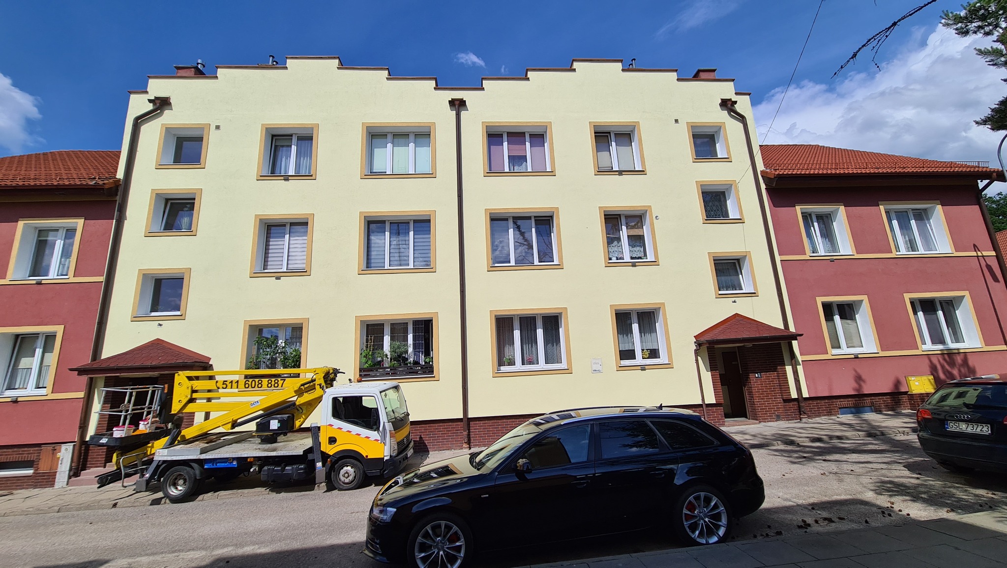 Żółta elewacja trzypiętrowego budynku z białymi oknami, widoczny żółty podnośnik na samochodzie dostawczym i czarny samochód osobowy na pierwszym planie.
