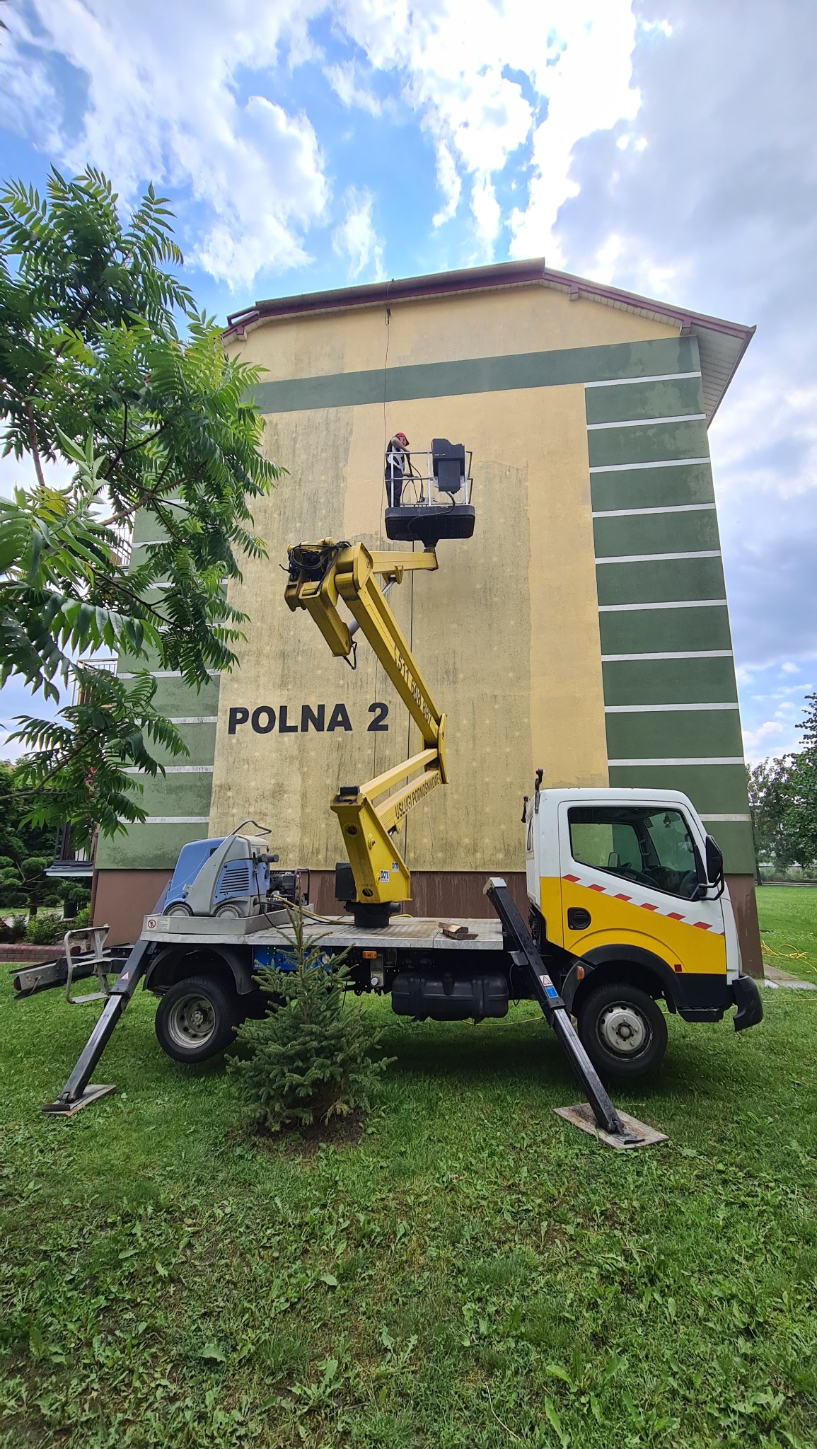 Pracownik w czerwonym kasku w koszu ruchomego podnośnika na tle budynku z napisem POLNA 2; żółty wysięgnik, biało-żółta ciężarówka na trawniku.