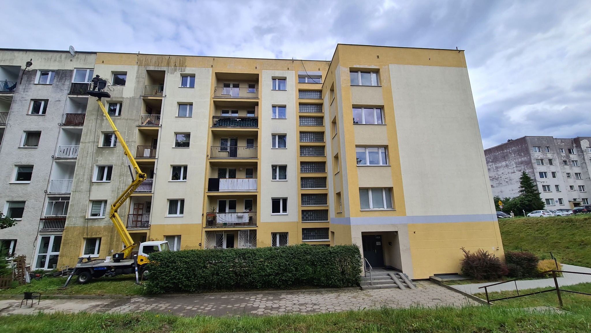 Żółty podnośnik koszowy z operatorem, na tle częściowo umytej elewacji bloku mieszkalnego, ukazujący różnicę w kolorze przed i po zabiegu.