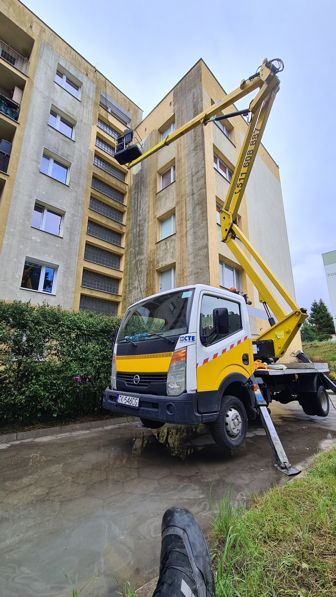 Żółty podnośnik koszowy na tle bloku z widocznymi śladami czyszczenia elewacji; mokry chodnik i fragment buta w dolnej części kadru.