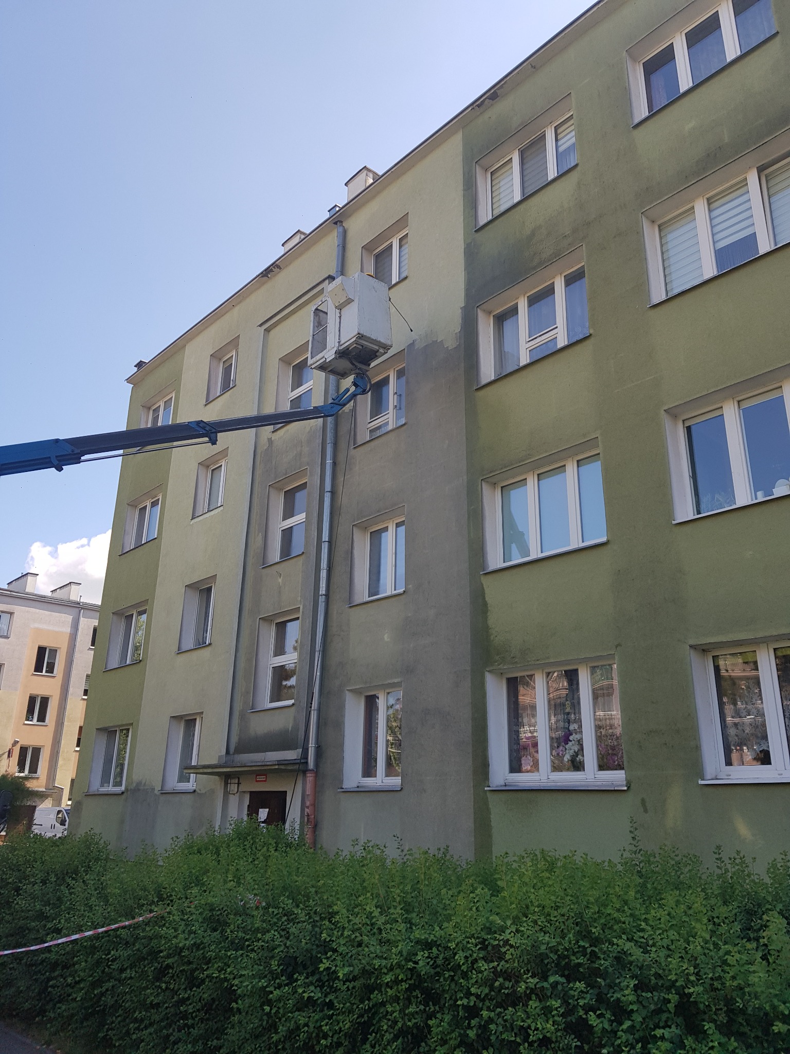 Fragment bloku mieszkalnego z widocznymi śladami czyszczenia elewacji za pomocą podnośnika koszowego, z zaznaczoną taśmą strefą bezpieczeństwa przy zielonym żywopłocie.