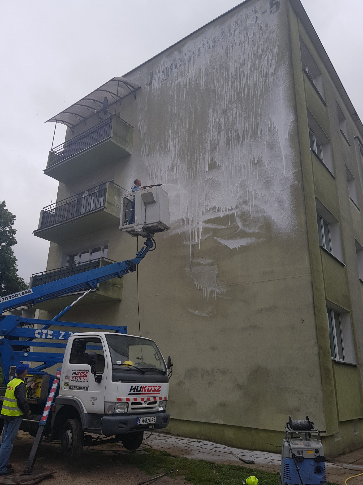Mycie elewacji budynku z podnośnika koszowego, widoczne ślady czyszczenia, pracownik w koszu, firma Hukosz i jej pojazd na dole
