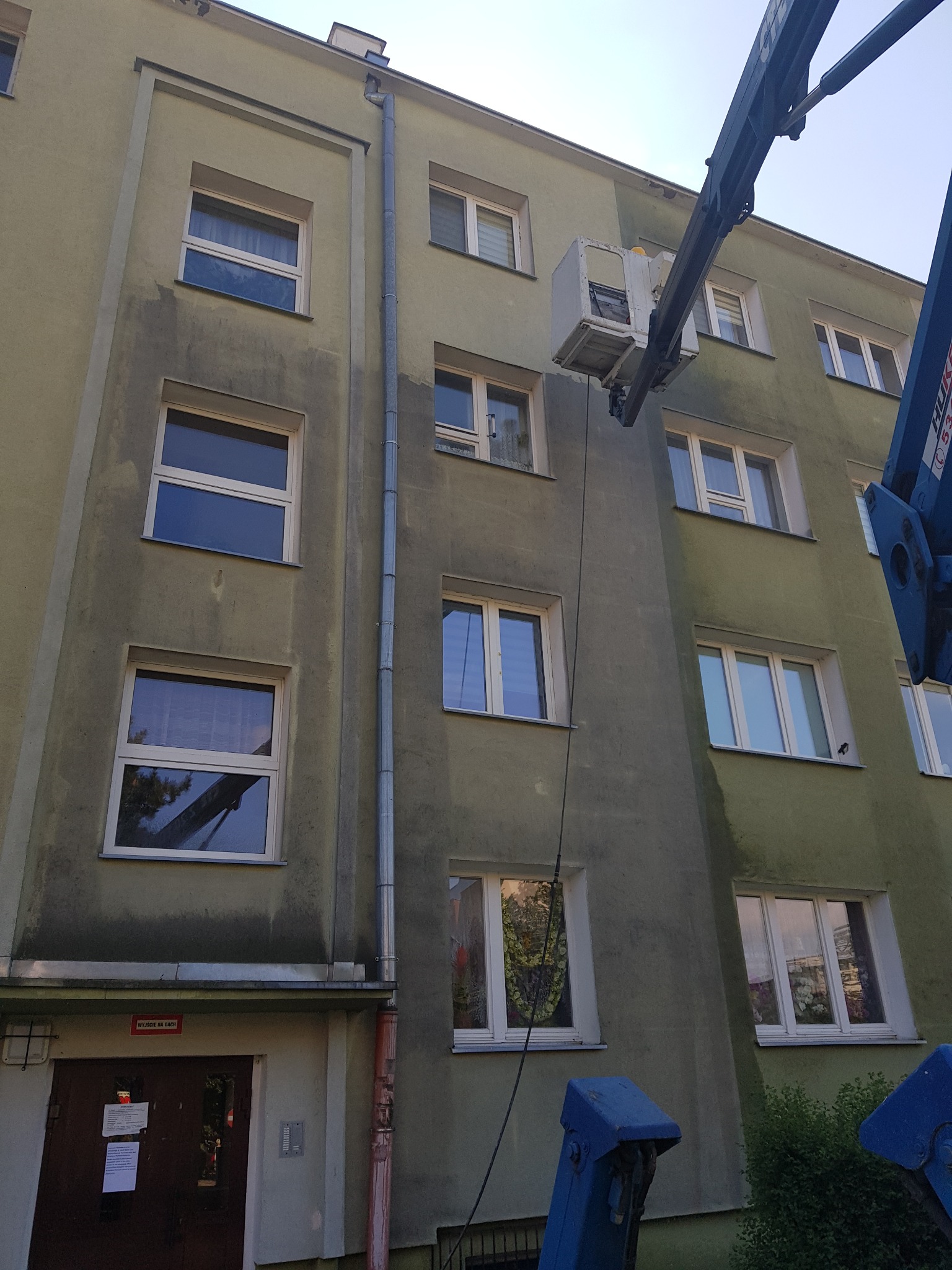 Mycie elewacji bloku z podnośnika koszowego, widoczne różnice w czystości ściany przed i po myciu, część kosza podnośnika w kadrze.