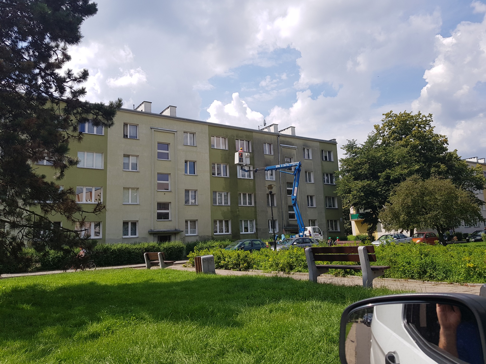 Pracownik w pomarańczowym kasku na podnośniku koszowym podczas prac na elewacji budynku wielorodzinnego, widok zza drzewa i fragmentu lusterka samochodu.