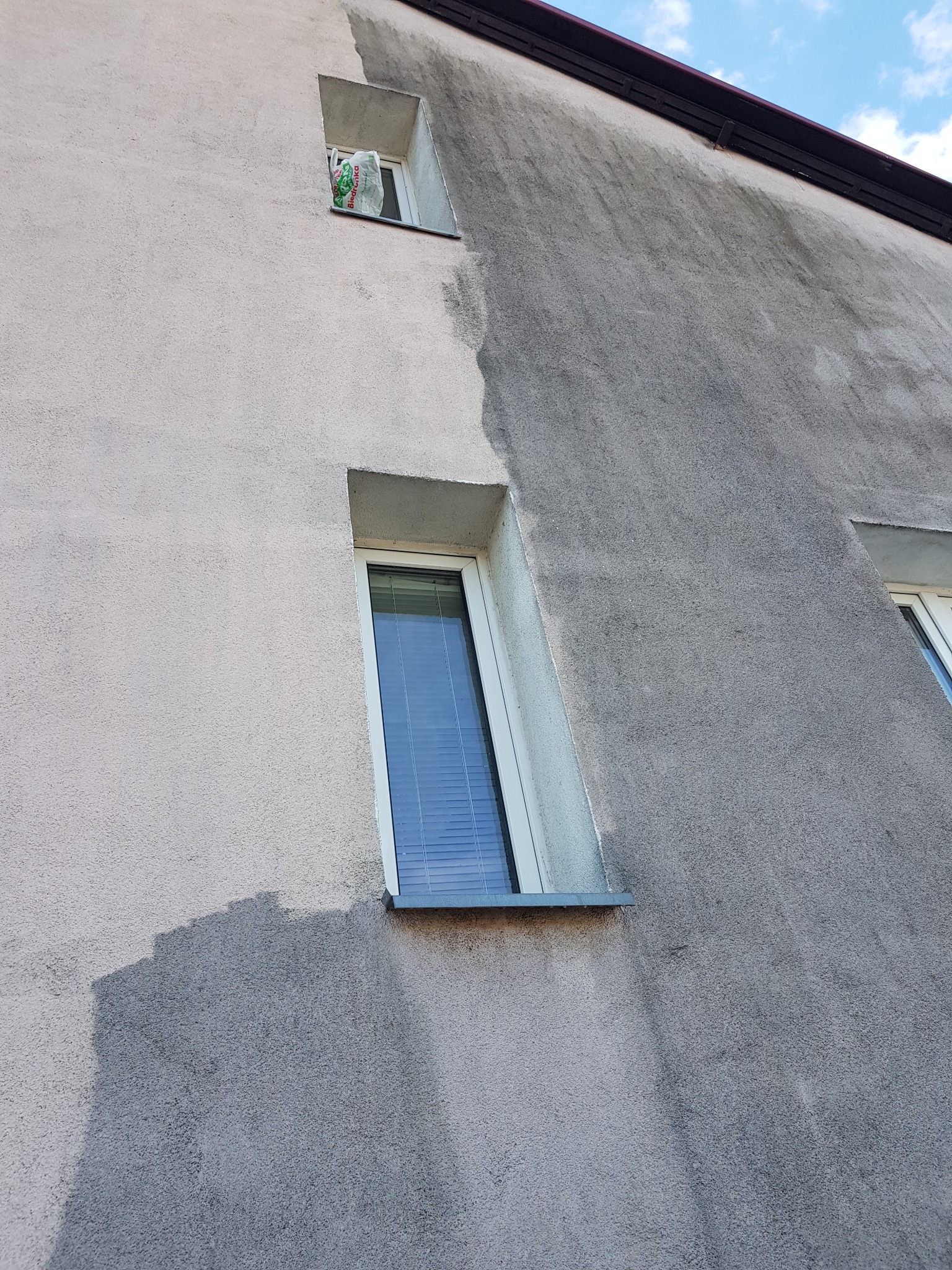 Częściowo umyta elewacja budynku z oknami, widoczna wyraźna różnica między czystą a brudną powierzchnią, na parapecie jednego z okien torba z Biedronki.