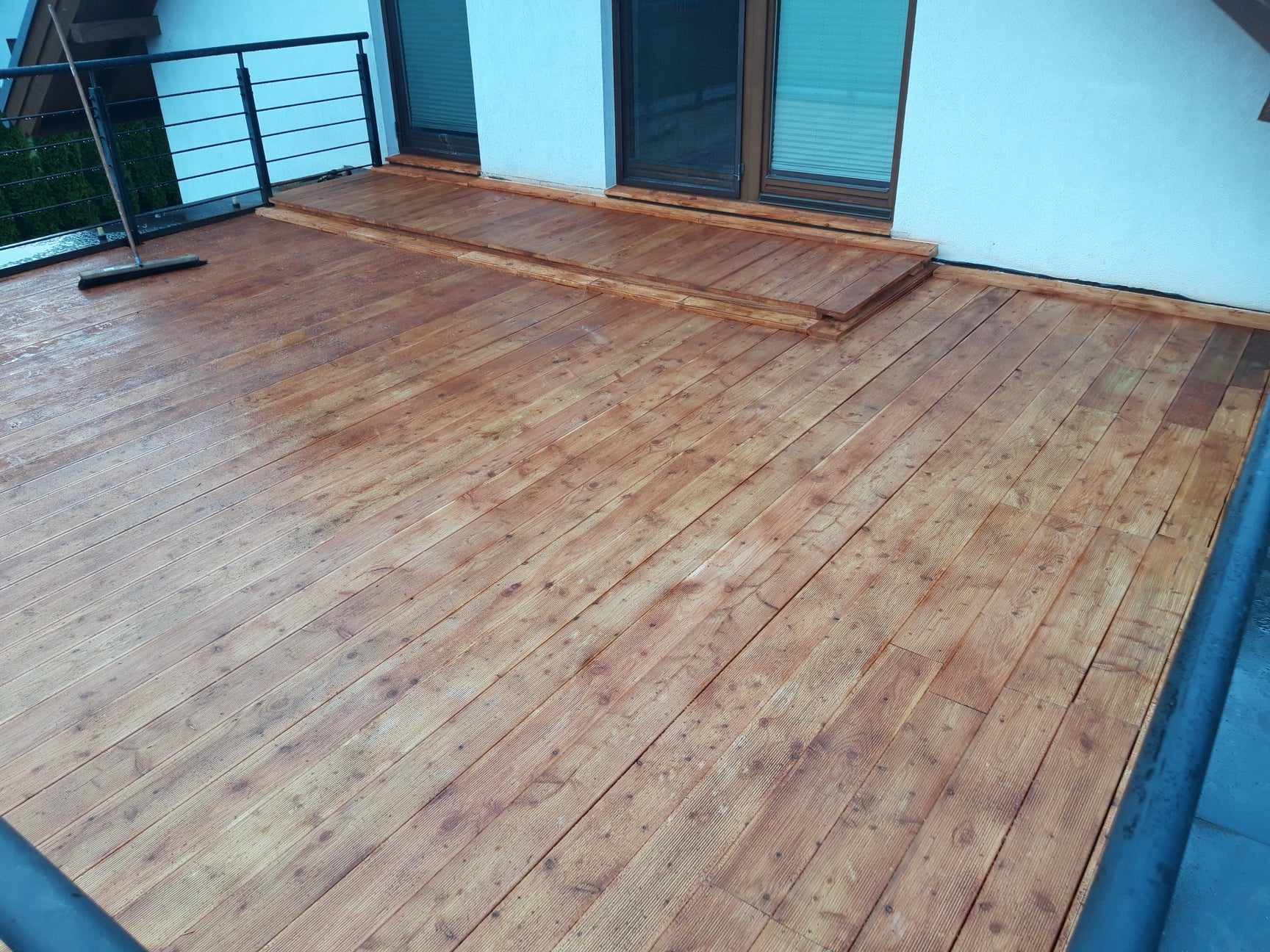 Balkon z drewnianą podłogą, świeżo po umyciu, z widoczną mokrą miotłą opartą o balustradę.