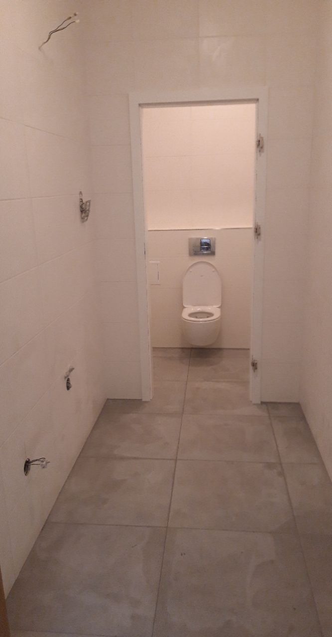 Wnętrze łazienki w trakcie wykańczania, widoczne szare płytki podłogowe, białe płytki ścienne, wisząca toaleta i wystające kable ze ściany.