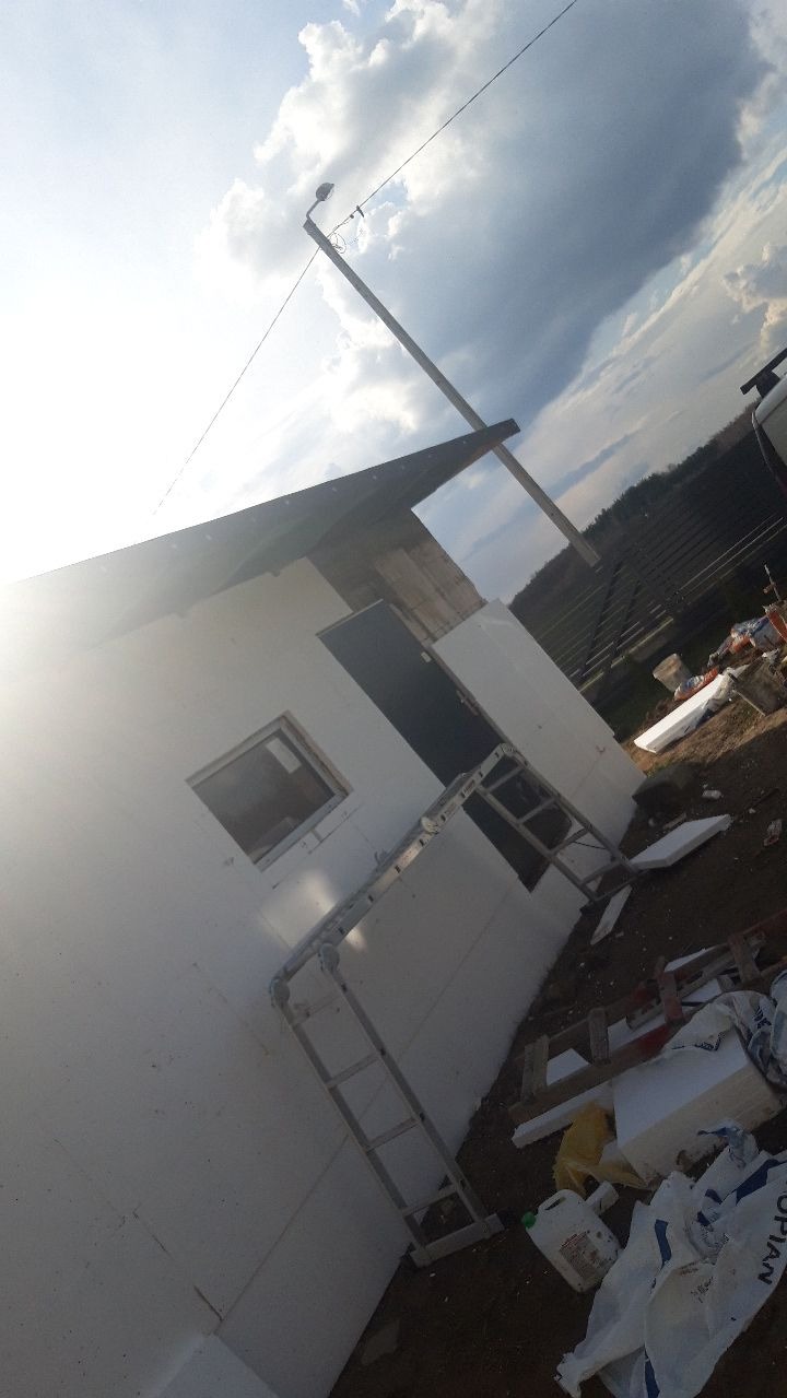 Budowa małego domu z ociepleniem styropianowym, widoczna drabina oparta o ścianę, fragment dachu i niebo z chmurami w tle.