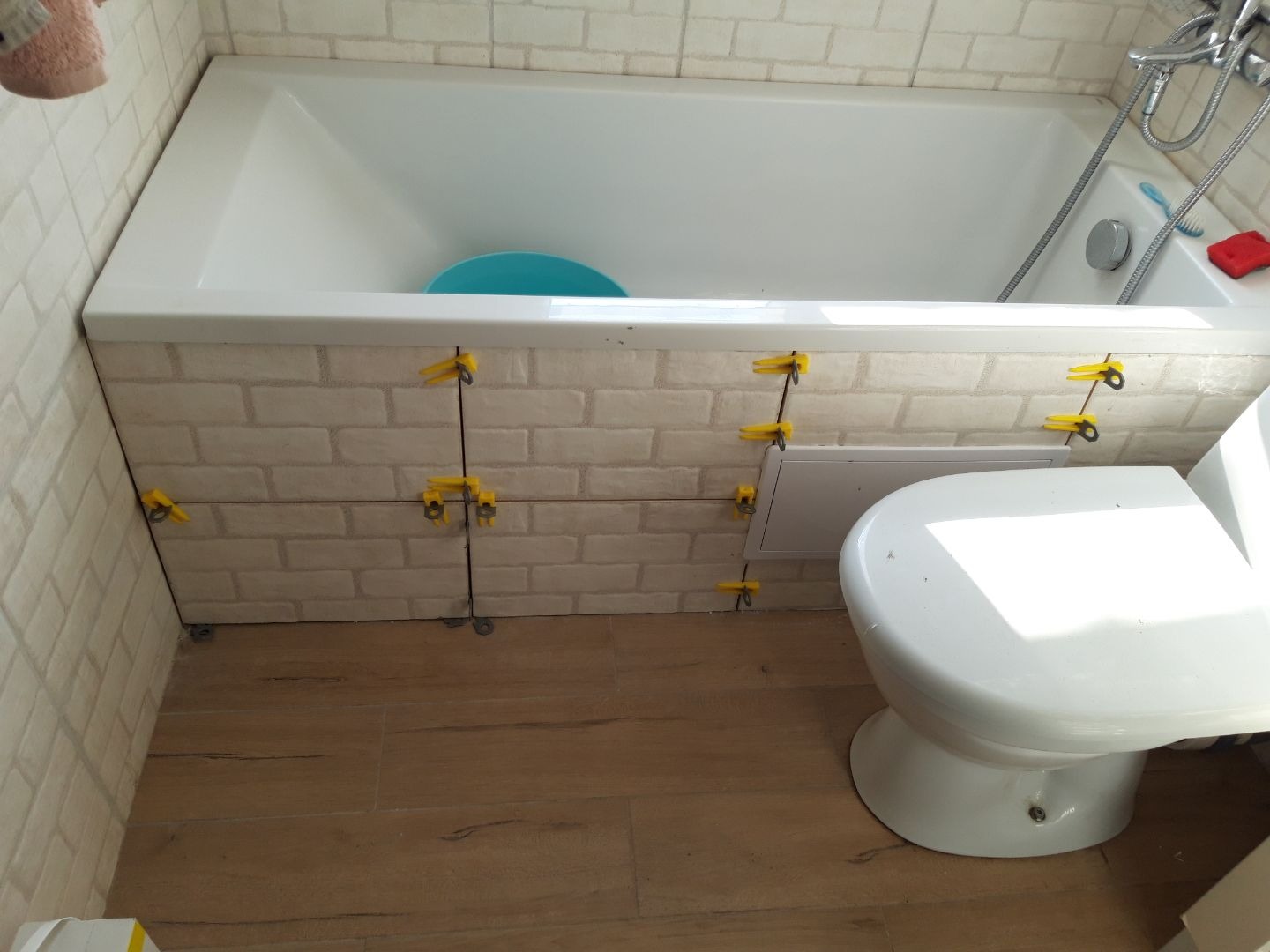 Świeżo wykafelkowana obudowa wanny w łazience, z zastosowaniem poziomowania płytek za pomocą klipsów i klinów, widoczny również fragment toalety i drewnopodobnej podłogi.