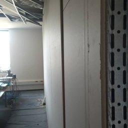 Maciej Stawiarski - Montaż płyty kartonowo-gipsowej na metalowym stelażu w pomieszczeniu biurowym, widoczne przewody wentylacyjne pod sufitem i zabezpieczona folią podłoga.