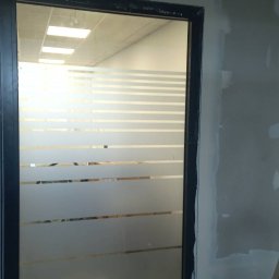 Maciej Stawiarski - Ściana z niedokończonymi pracami wykończeniowymi, widoczna przez przeszklone drzwi z poziomymi paskami, na podłodze zabrudzenia po pracach budowlanych.