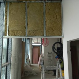 Maciej Stawiarski - Montaż izolacji z wełny mineralnej w metalowym stelażu sufitu podwieszanego, widoczne kable, drabina i pracownik w tle.