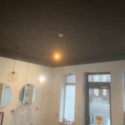 Maciej Stawiarski - Pomieszczenie w trakcie prac wykończeniowych: pomalowany na czarno sufit, białe ściany z okrągłymi lustrami i wiszącymi lampami, okna i drzwi zabezpieczone folią, widoczne ślady po instalacjach...