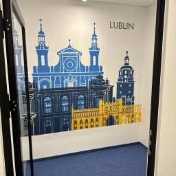Maciej Stawiarski - Wnętrze biura z dekoracyjną grafiką na ścianie przedstawiającą panoramę Lublina w kolorach niebieskim i żółtym, widoczne przez otwarte drzwi z czarną ramą.