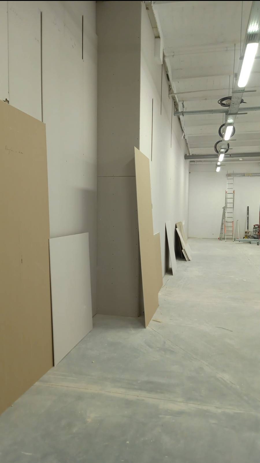 Montaż płyt karton-gips na ścianach w surowym wnętrzu, widoczne płyty przy ścianach i na podłodze, oświetlenie przemysłowe na suficie, w tle drabina i narzędzia.