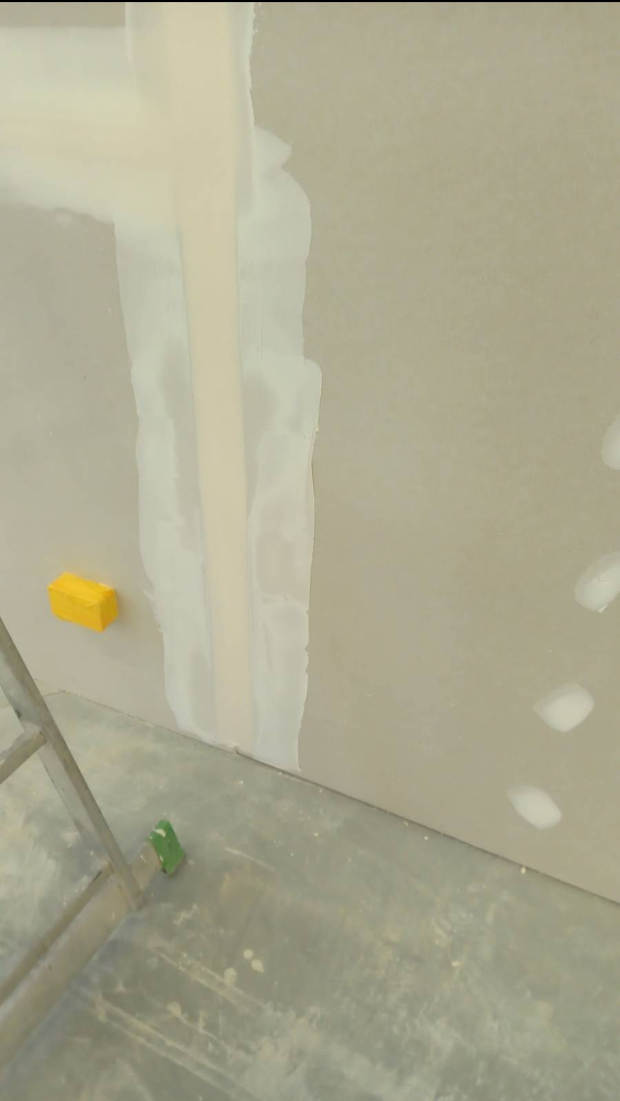 Wypełniona masa szpachlowa na łączeniu płyt karton gips z żółtym bloczkiem szlifierskim i fragmentem drabiny na podłodze.