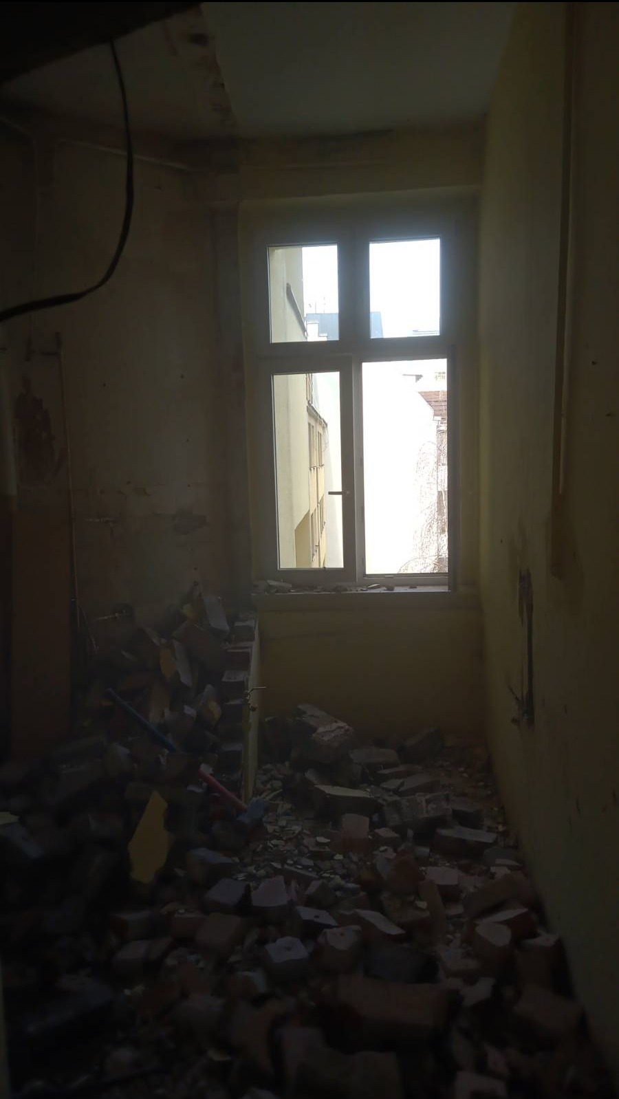 Pokój w trakcie demontażu, z gruzem ceglanym na podłodze i widocznym oknem z widokiem na budynek sąsiedni.