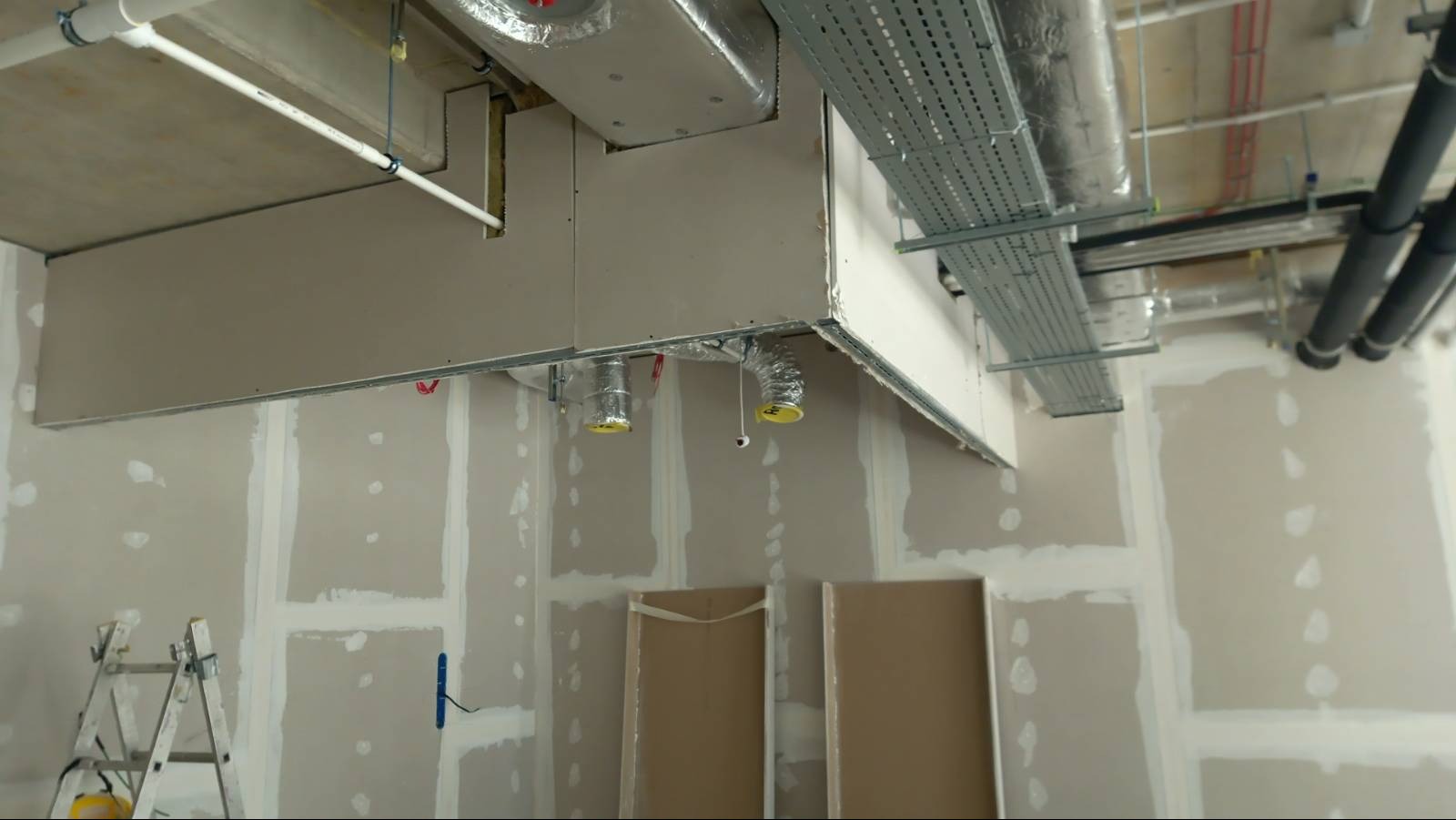 Montaż instalacji wentylacyjnej pod sufitem podwieszanym z płyt kartonowo-gipsowych, widoczne przewody wentylacyjne, drabina i szpachlowane ściany w trakcie prac wykończeniowych.