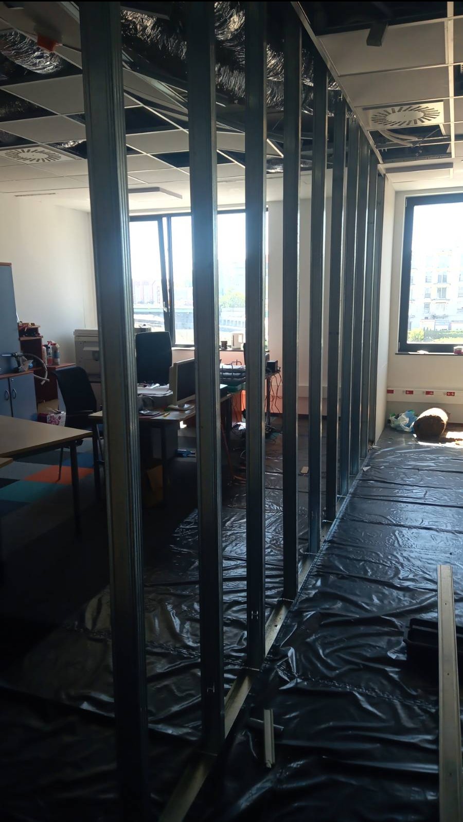 Stelaż z profili stalowych do ścianki działowej z płyt kartonowo-gipsowych w biurze, zabezpieczony folią ochronną na podłodze, widoczne okno i biurka w tle.