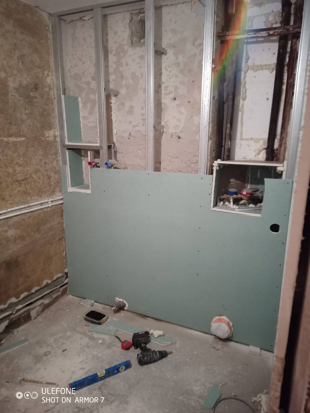 Montaż płyty gipsowo-kartonowej w łazience, widoczne stalowe profile, otwory na instalacje wodne, poziomica i wiertarka na podłodze, surowy stan wykończenia.