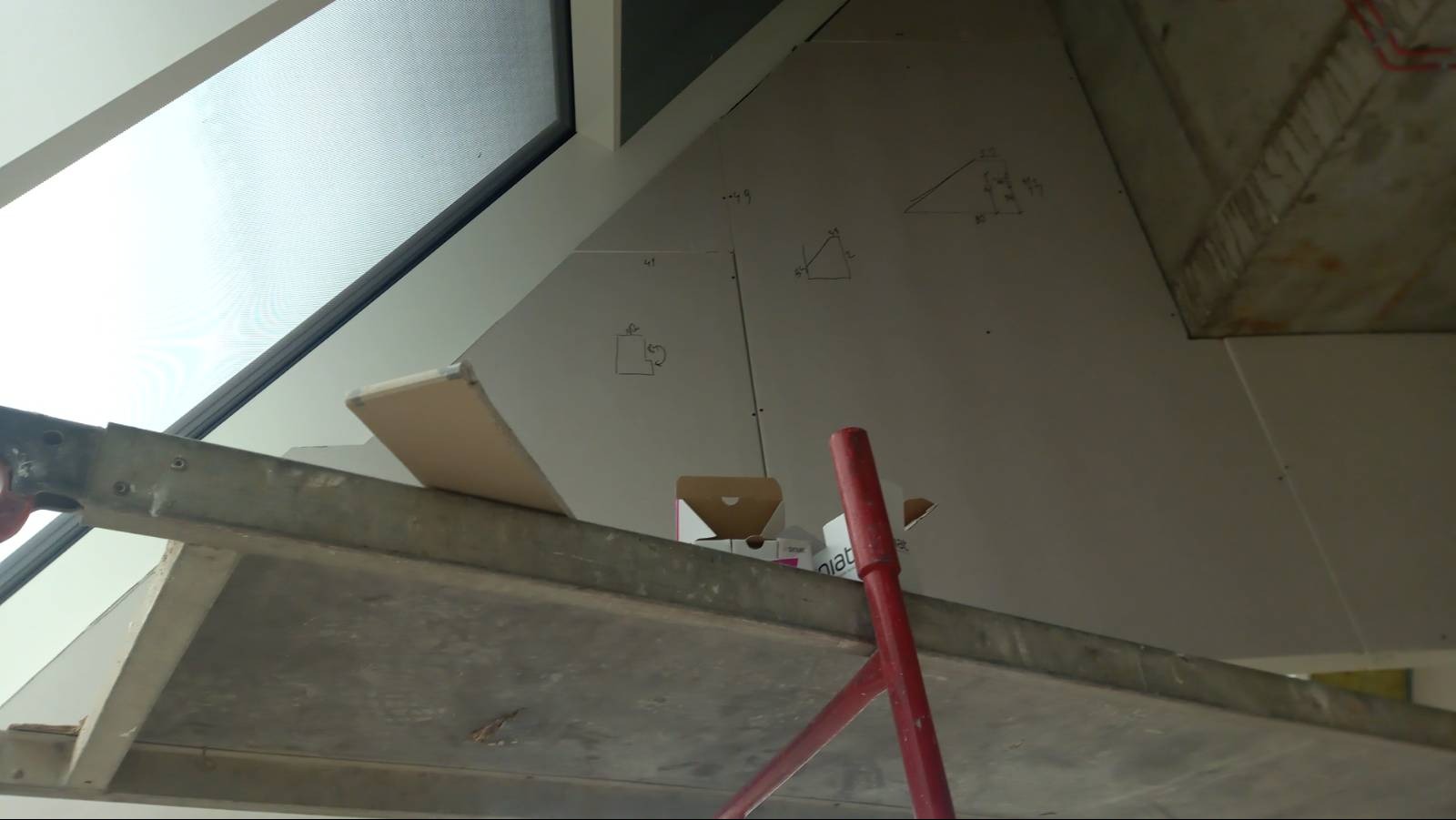 Montaż płyty gipsowo-kartonowej na poddaszu, widoczne rusztowanie, okno dachowe i odręczne szkice z wymiarami na płycie.