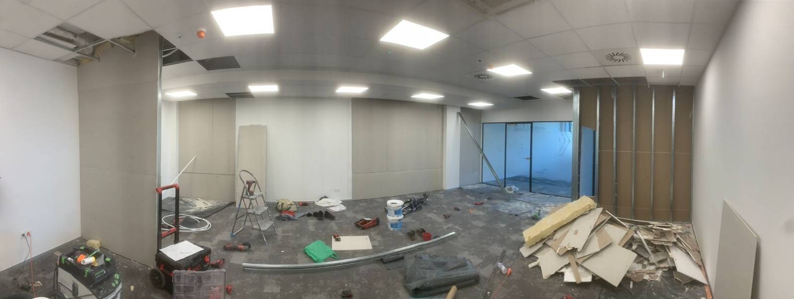 Wnętrze biurowe w trakcie montażu ścian działowych z płyt kartonowo-gipsowych na metalowej konstrukcji, widoczne narzędzia, drabina, płyty gipsowe i pozostałości po pracy na podłodze.