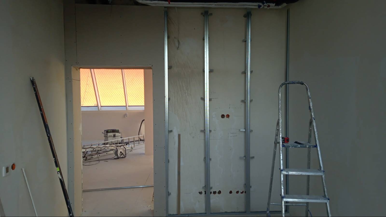 Montaż metalowego stelażu pod płyty kartonowo-gipsowe na ścianie, z widoczną drabiną i narzędziami, oraz fragmentem pomieszczenia z oknem dachowym w tle.