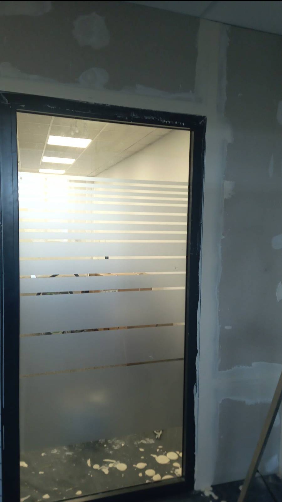 Ściana z niedokończonymi pracami wykończeniowymi, widoczna przez przeszklone drzwi z poziomymi paskami, na podłodze zabrudzenia po pracach budowlanych.