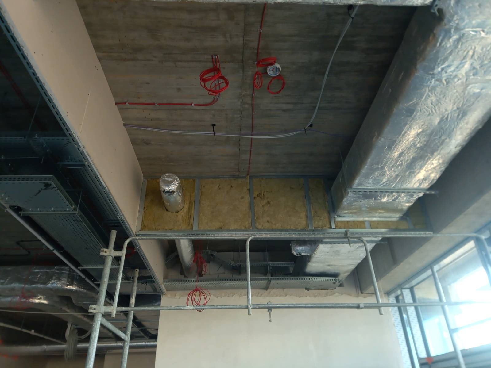 Montaż izolacji z wełny mineralnej w metalowym stelażu pod sufitem, widoczne kable elektryczne i rury wentylacyjne, ujęcie z rusztowania.