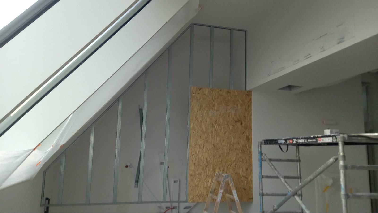 Montaż metalowego stelaża pod płytę gipsową na poddaszu ze skośnym oknem dachowym, widoczna płyta OSB, drabina i rusztowanie malarskie.