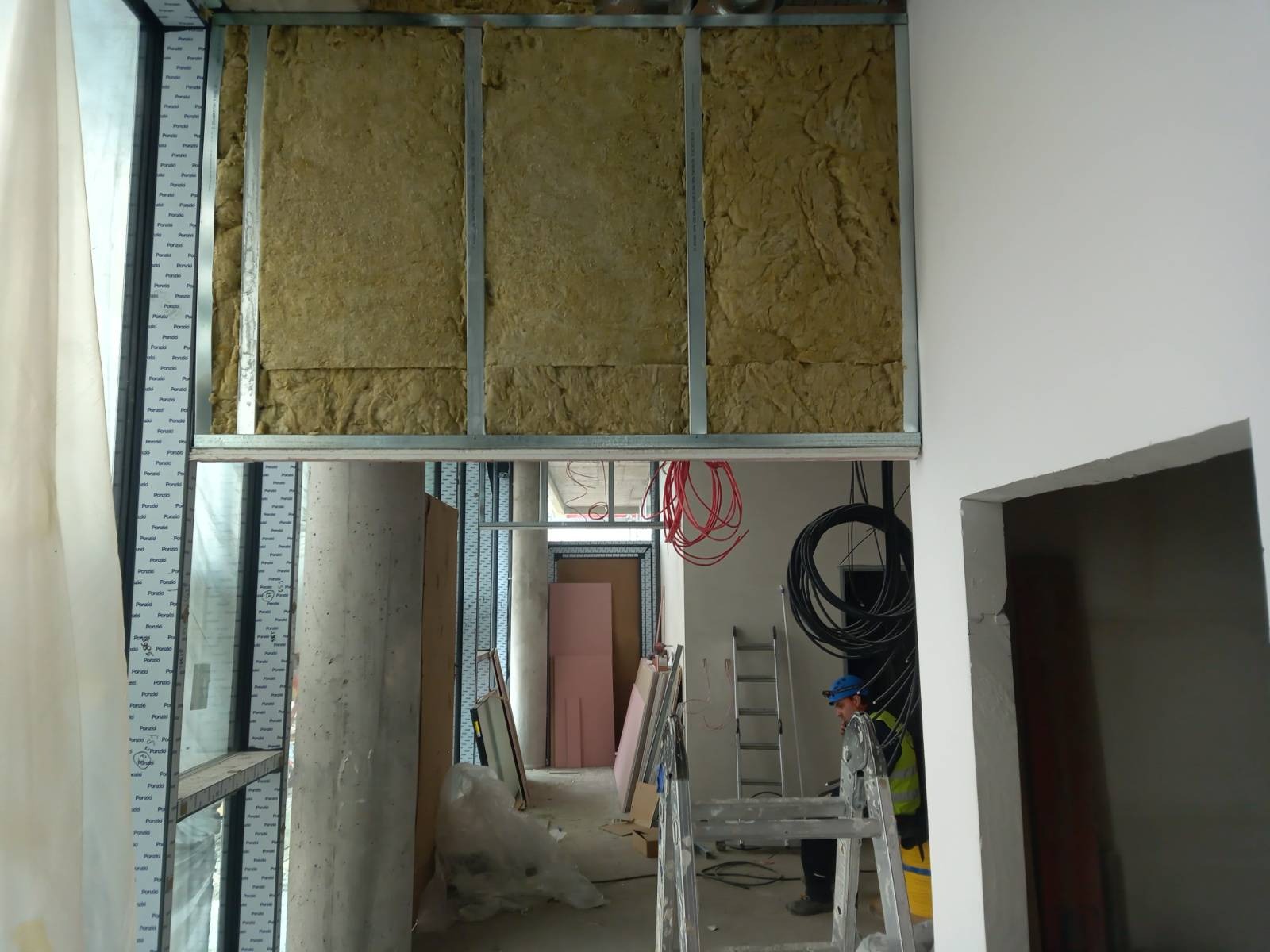 Montaż izolacji z wełny mineralnej w metalowym stelażu sufitu podwieszanego, widoczne kable, drabina i pracownik w tle.