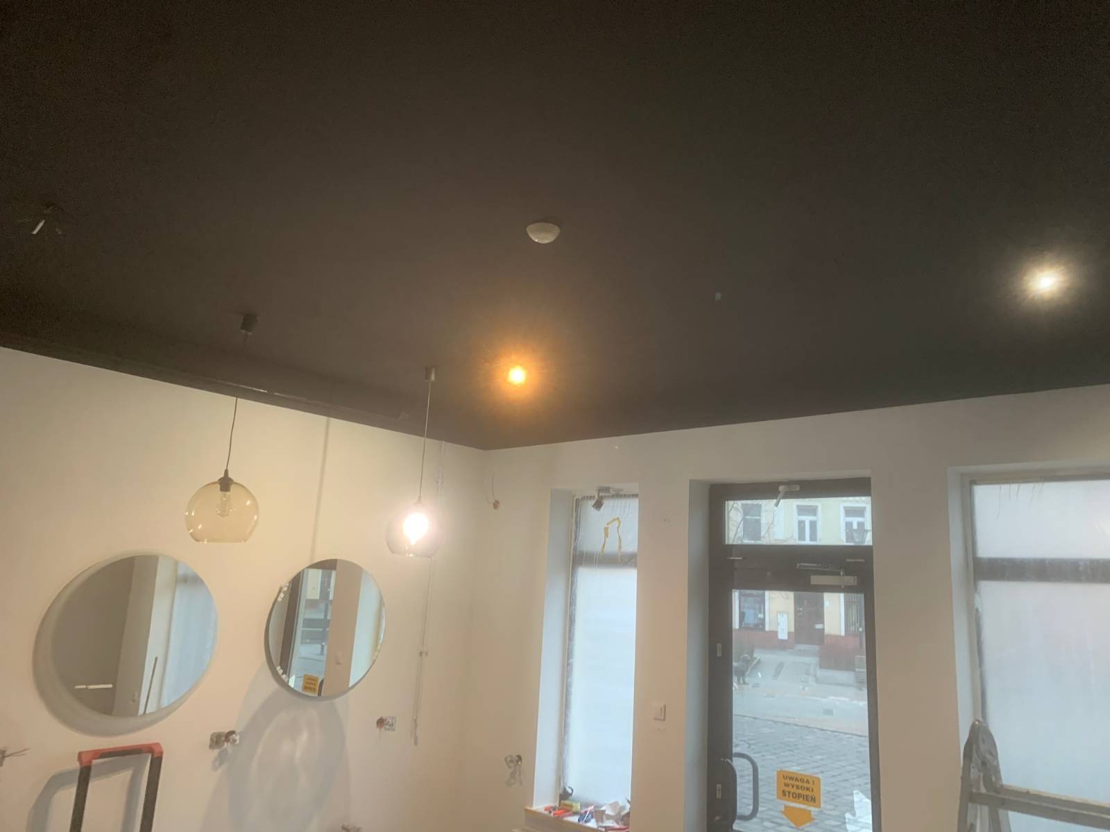 Pomieszczenie w trakcie prac wykończeniowych: pomalowany na czarno sufit, białe ściany z okrągłymi lustrami i wiszącymi lampami, okna i drzwi zabezpieczone folią, widoczne ślady po instalacjach...