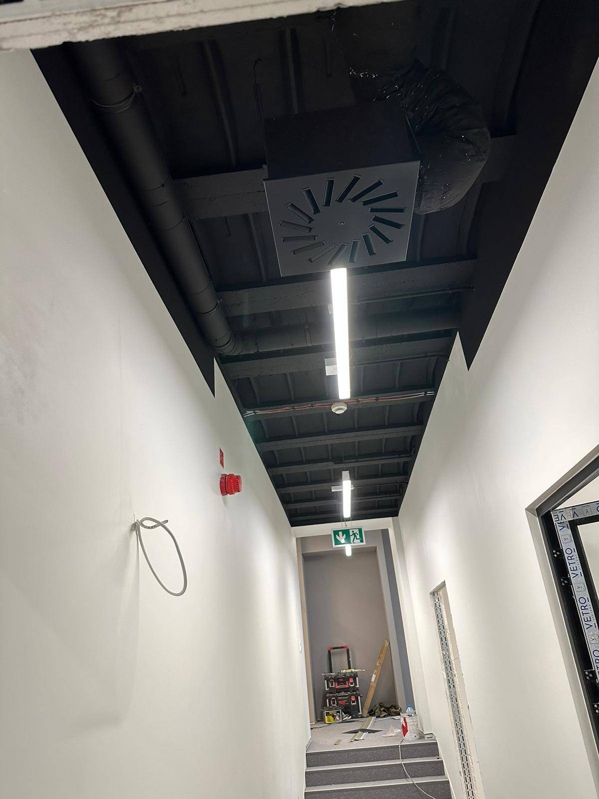 Korytarz w trakcie wykańczania: pomalowany sufit na czarno z widoczną instalacją wentylacyjną, białe ściany z elementami instalacji przeciwpożarowej i oświetleniem awaryjnym, narzędzia na podłodze.